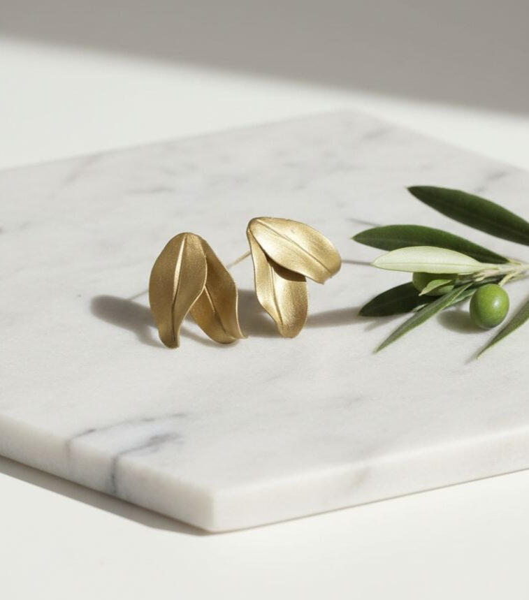 Pendientes de botón con hojas de olivo en porcelana fría color oro envejecido, colocados sobre una superficie de mármol junto a una ramita de olivo, diseño artesanal de Pajarita Presumida.